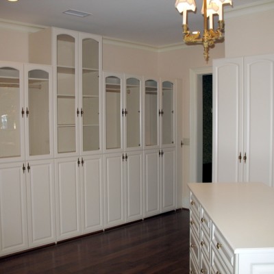 Cabinets deep enough for hanging clothes behind arched top glass doors / solid raised panel doors are punctuated by a 10' tall middle shelf unit. Antique white laminates offer a subdued look in a large dressing room.